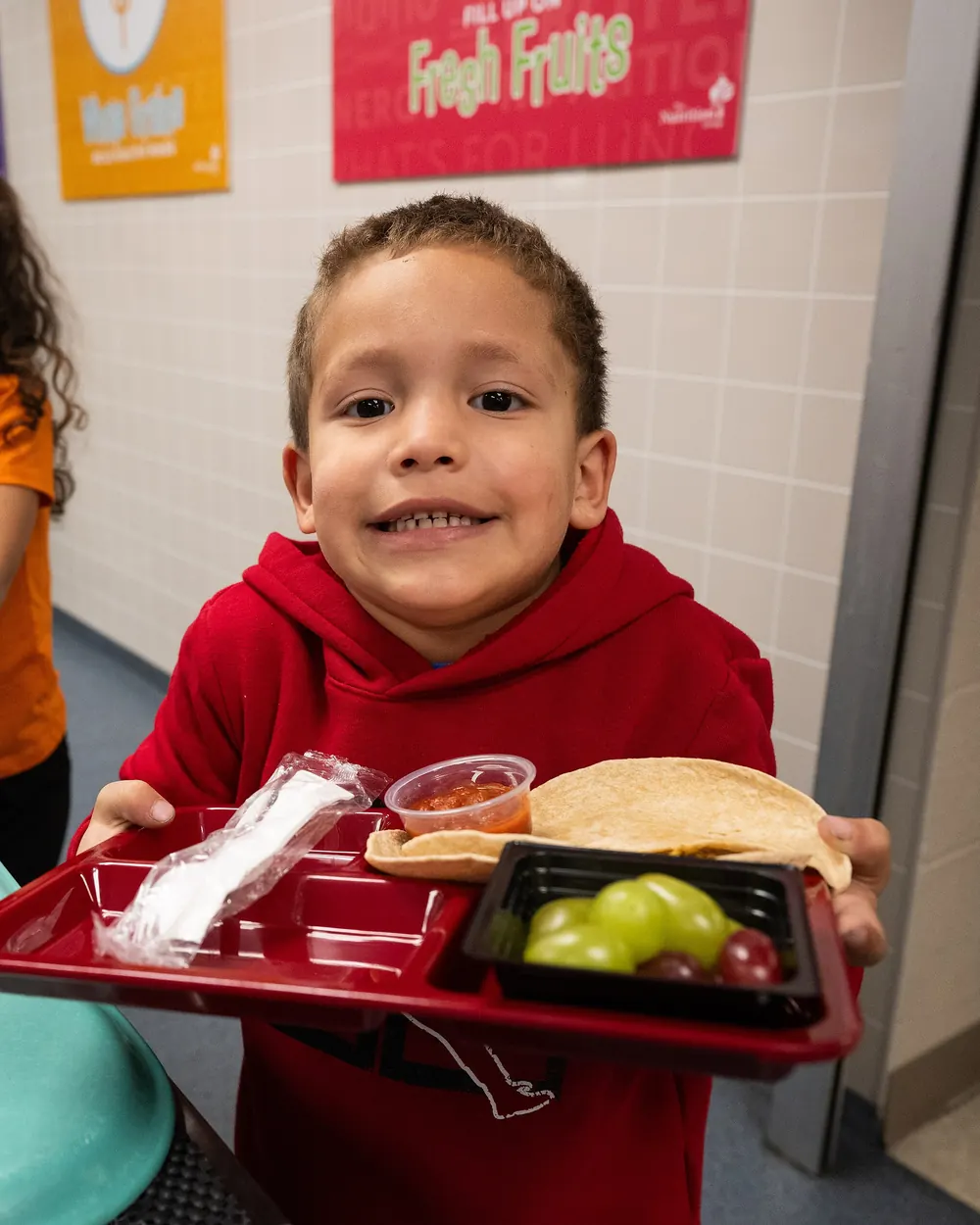 A smiling boy holds up his lunch tray in a school cafeteria.